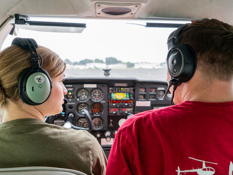 Two people wearing headsets sit in a small aircraft cockpit, looking at the control panel. The mood is focused and cooperative.