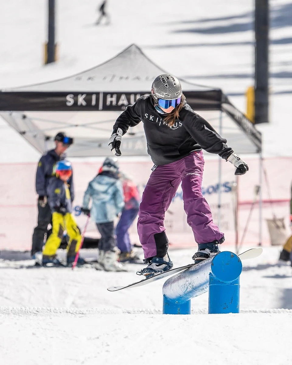 Snowboarder jumpin in front of Ski Haus branded canopy at a winter sports event