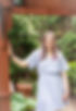 Image of a woman with light purple dyed hair and a light blue dress on. The woman is in a garden and is smilng. One hand is by her side and the other is on a brown archway.
