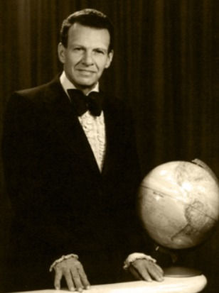 Retrato de um homem na década de 1950, vestindo smoking com gravata borboleta, segurando uma mesa com um globo terrestre ao lado, em um fundo de cortinas com aspecto clássico.
