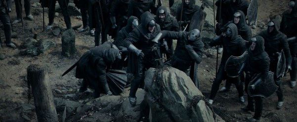 Grupo de guerreiros vestidos de preto, com capuzes e armaduras, reunidos em um terreno rochoso e sombrio, alguns empunhando escudos e armas.