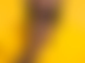 Homem negro com barba vestindo uma camiseta amarela, fazendo o gesto de silêncio com o dedo indicador sobre os lábios, sobre um fundo amarelo.