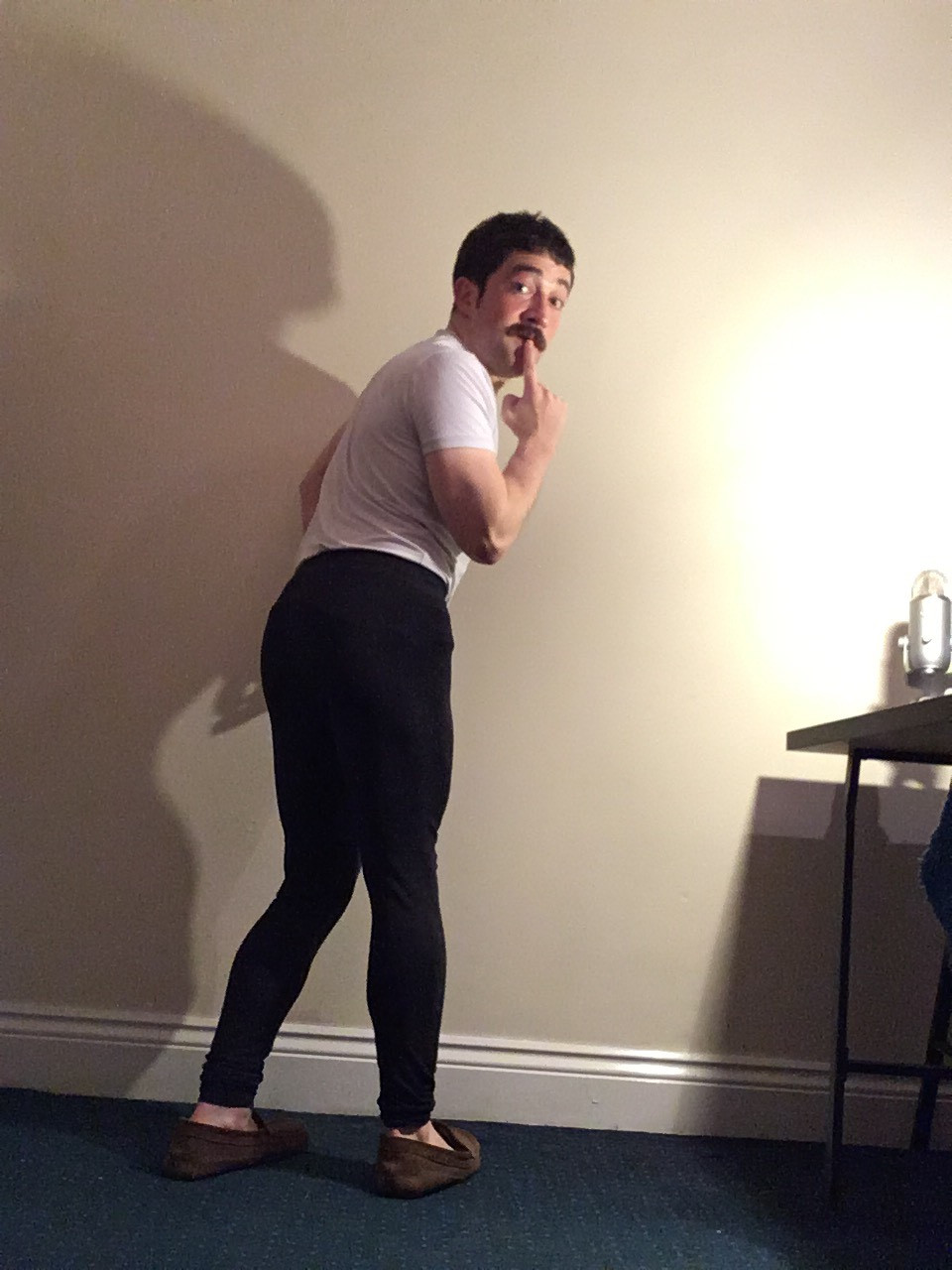 A white man with a moustache makes a comedic coquettish face as he looks over his shoulder
