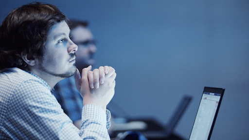 Man in checkered shirt listens thoughtfully in a blue-lit meeting room, hands clasped, with a laptop and blurred colleague behind.