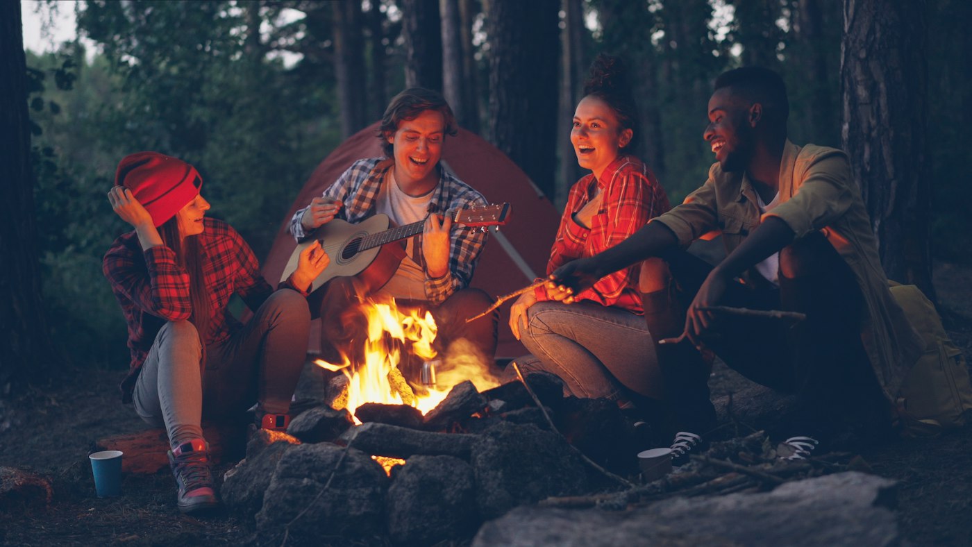 Real editorial photograph of friends laughing around a campfire. Cover for A Potluck Is a Form of Shared Culture