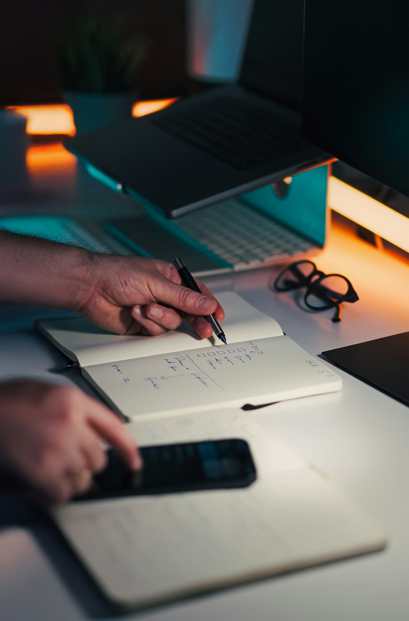 Real editorial photograph of hands writing next to laptop and phone. Cover for Your Own Tiny Habit Experiment