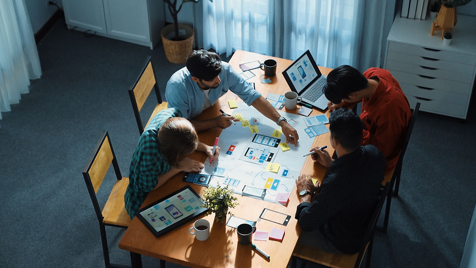 Four people collaborate at a table with laptops, tablets, and design sketches. Bright room with blue curtains. Engaged in teamwork.