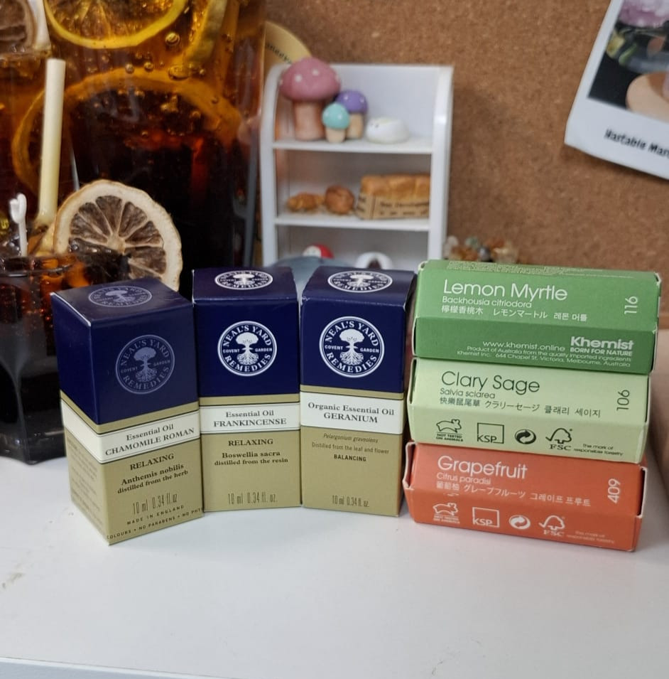 Neal's Yard Remedies and Khemist essential oil boxes displayed on a workspace table next to a decorative drink and miniature model.