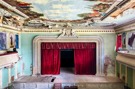 An abandoned and derilict, but beautiful, colourful theatre with a half-open red satin curtain, a church-like ceiling with paintings and a wooden stage with holes in it.