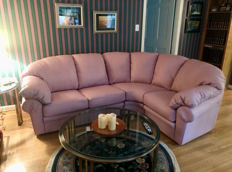  Large curved lilac-colored sectional sofa in a living room with striped green and pink wallpaper, a round glass coffee table with candles, and a floral rug, representing comfortable seating options from Mill End Shops Rockville MD