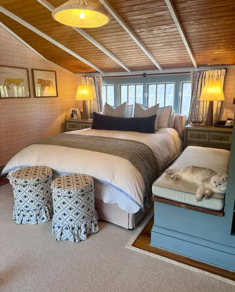 ozy bedroom with a large bed, two patterned ottomans at the foot of the bed, a built-in bench with a cat lounging on it, and windows with drapes, showcasing bedroom design and soft furnishings from Mill End Shops Fairfax VA