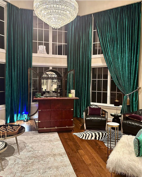 Luxurious living room featuring tall emerald green velvet curtains on large windows, a large crystal chandelier, a vintage bar cabinet, and leather armchairs with a zebra print rug, displaying dramatic window treatments from Mill End Shops Rockville MD