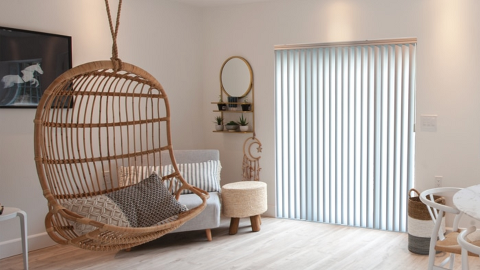 White Synchrony™ vertical blinds by Norman® USA in a cozy, minimalist living room with a rattan hanging chair and neutral furniture.