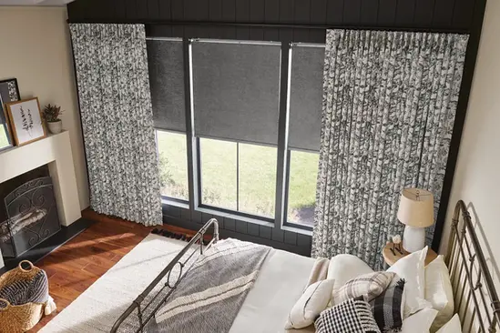 Bedroom view with blackout roller shades in slate gray, layered under floral artisan pleated drapes.