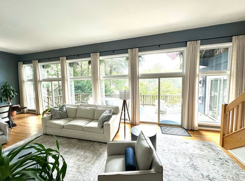 Spacious living room with a wall of large windows and sliding glass doors leading to a deck, featuring light-colored custom drapes, a cream sofa, and a light-colored rug, illustrating large window treatments from Mill End Shops Fairfax VA