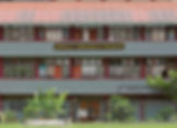 Two-story school building with "Sekolah Kebangsaan Bangsar" sign. Red roof, fenced yard with plants, and visible posters on windows.