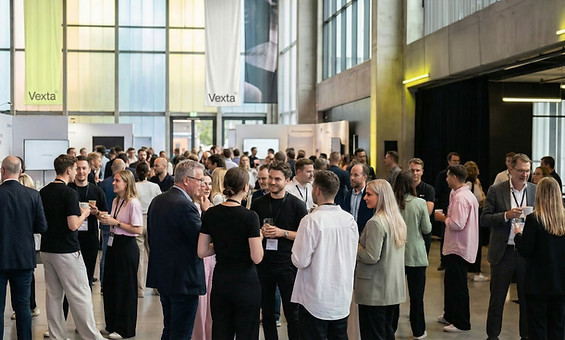 The setting is a foyer of a large building, maybe a gallery or function space, with mixed groups of adults standing, drinking & chatting.