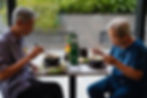 Two elderly individuals enjoy noodle soup at a cafe, seated at PHO24. Lush greenery is outside the window, creating a peaceful atmosphere.