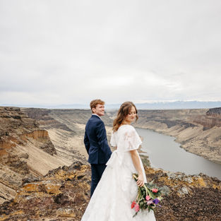 Couple facing away looking back at the camera. Bride is in wedding dress man behind her in suit standing in front of Jordanelle Reservoir.