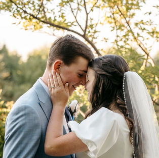 Bride and groom forehead to forehead in front of some trees. Groom is wearing a light gray suit. Brides dress has puff sleeves.