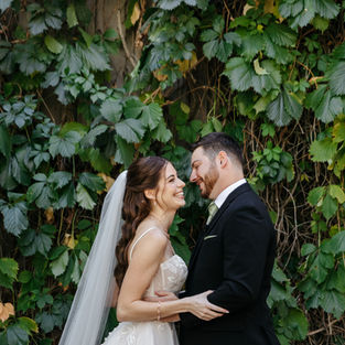 Bride and groom standing in front of a green hedge smiling and laughing together. Groom is wearing a black suit and the bride has a long veil.