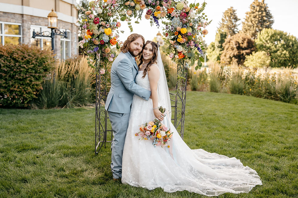 A couple embraces under a colorful floral arch in a garden. The bride holds a bouquet, wearing a white gown; the groom is in a light blue suit.