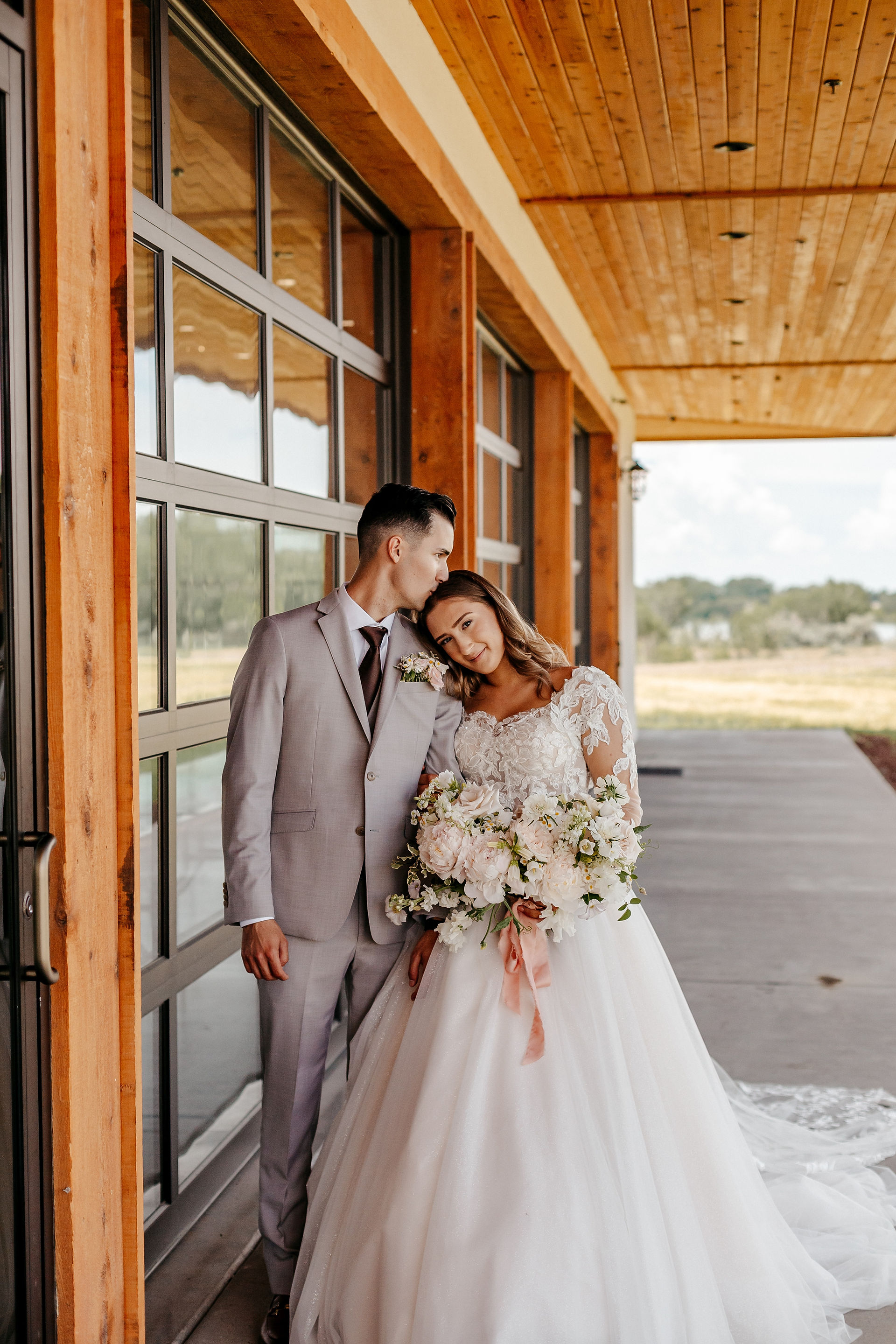 Wedding couple standing together. The groom is kissing the bride on the forehead as she looks towards the wedding photographer.