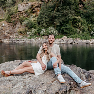 Engaged couple sitting together on a large rock in front of a mountain lake. Both smiling towards the camera.