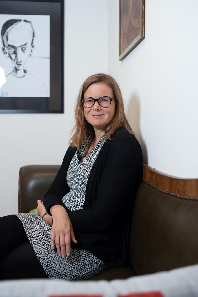 Caroline Depuydt sits on sofa hands folded and looks at the camera.
