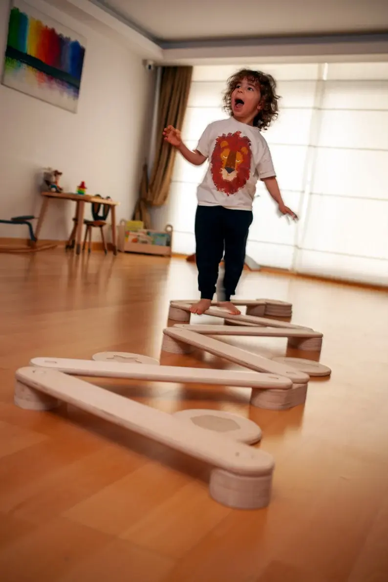 A toddler with curly hair walking on a wooden balance beam in his play room.