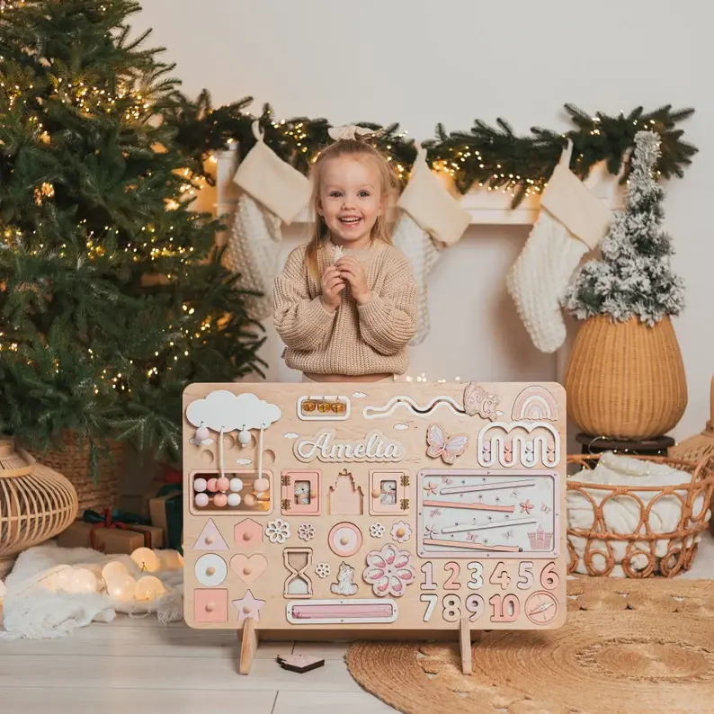A little girl standing behind her wooden busy board by the christmas tree.