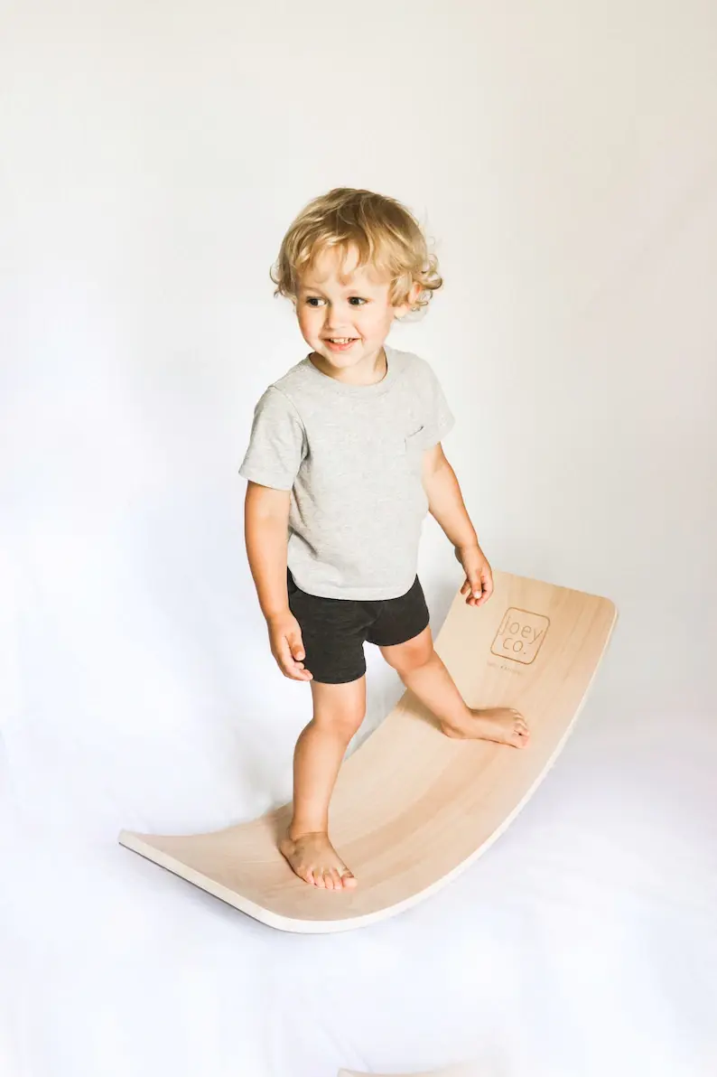 A blonde haired boy balancing on a wooden wobble board.