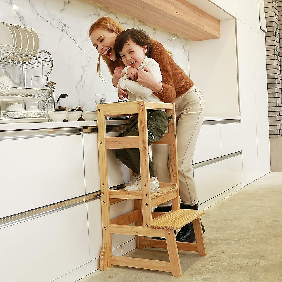 A mom laughing with her toddler boy who is standing on a kitchen stool in their modern white kitchen.