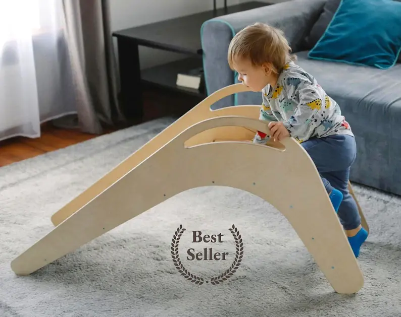 A toddler boy climbing up a wooden slide in his living room.