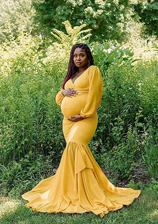 A woman in a yellow fitted maternity dress with one hand below and one hand above her baby bump in a green forest.