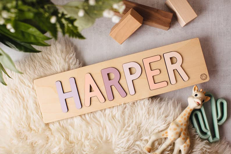 A wooden name puzzle for "HARPER" on the ground next to a rubber cactus, some stacking blocks, and a teether giraffe toy.