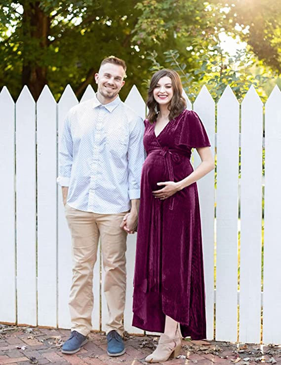 A pregnant woman in a burgundy maternity dress standing next to her husband in front of a white picket fence.
