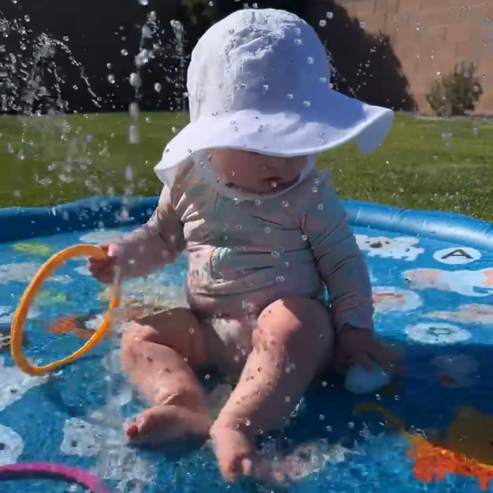 A baby girl sitting outside on a splash pad wearing a white sun hat.