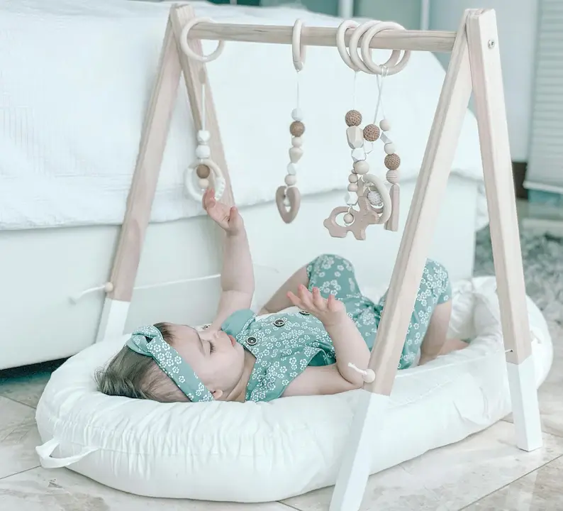 A baby in a green jumper laying underneath a wooden play gym in the living room.