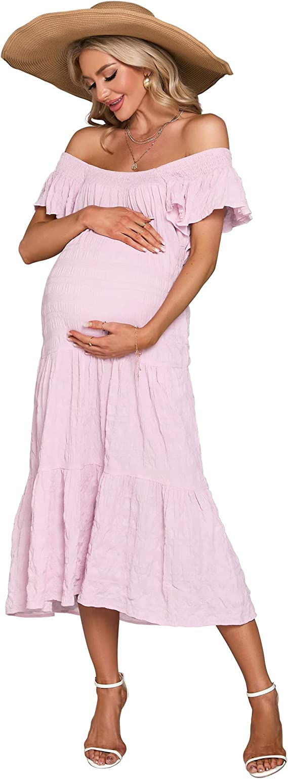 A blonde woman in a large brown hat and textured pink maternity dress holding her belly.