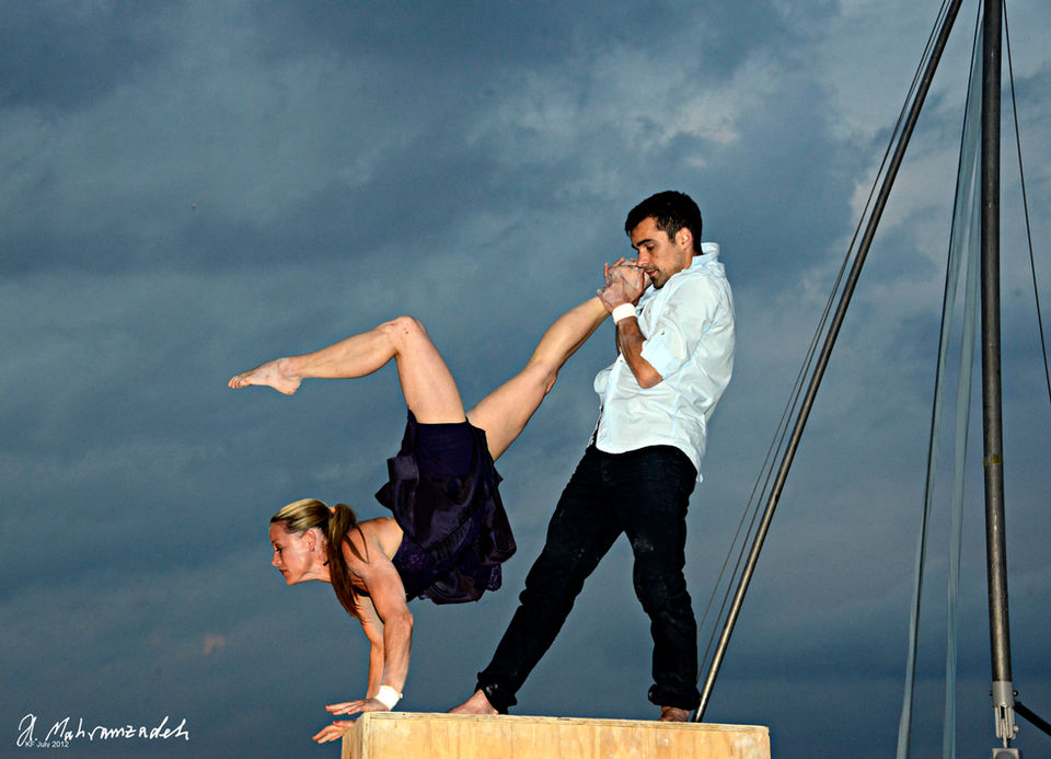 Een vrouwelijke acrobaat staat op haar handen terwijl een mannelijke acrobaat haar been vasthoudt op een houten blok. Een krachtige en elegante pose die de samenwerking van het duo benadrukt