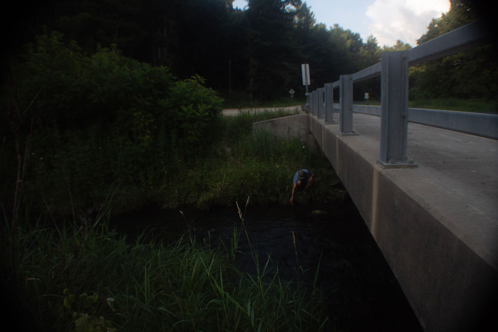 Person crouches by a creek under a gray roadside bridge, reaching into the water amid dark green woods.