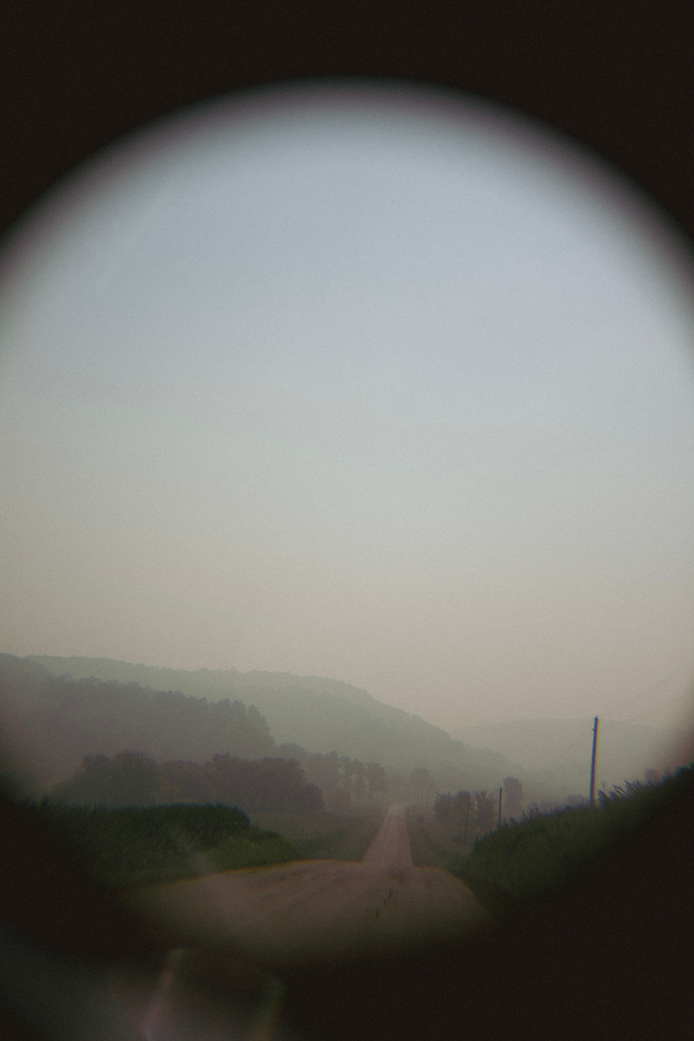 Blurred view through a circular lens of a misty rural road winding toward hazy hills, with dark trees and poles.