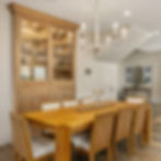 A traditional-style dining room featuring a chandelier hanging over a light ash wood table. Adjacent is a floor-to-ceiling oiled beech wood dining cabinet with two large open-frame doors, displaying decorative pieces brought to life by Kitchen and Bath Beyond.