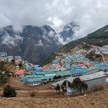 Mountain village with colorful rooftops under cloudy skies. Hills, scattered trees, and misty mountains in the background create a serene mood.