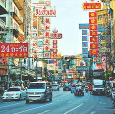 曼谷唐人街的街景，两旁建筑密集、招牌林立，以红色与金色为主，街道上车辆与嘟嘟车穿梭，行人在骑楼下活动，展现热闹的城市氛围。