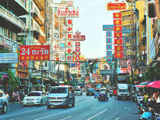 曼谷唐人街的街景,两旁建筑密集、招牌林立,以红色与金色为主,街道上车辆与嘟嘟车穿梭,行人在骑楼下活动,展现热闹的城市氛围。