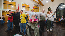 A diverse choir practices in a warmly lit room with wooden beams and flags. Members hold music sheets, focusing intently on singing.