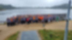 Group of people in gray and orange shirts raise hands, smiling by a lakeside with overcast skies and trees in the background. Joyful mood.