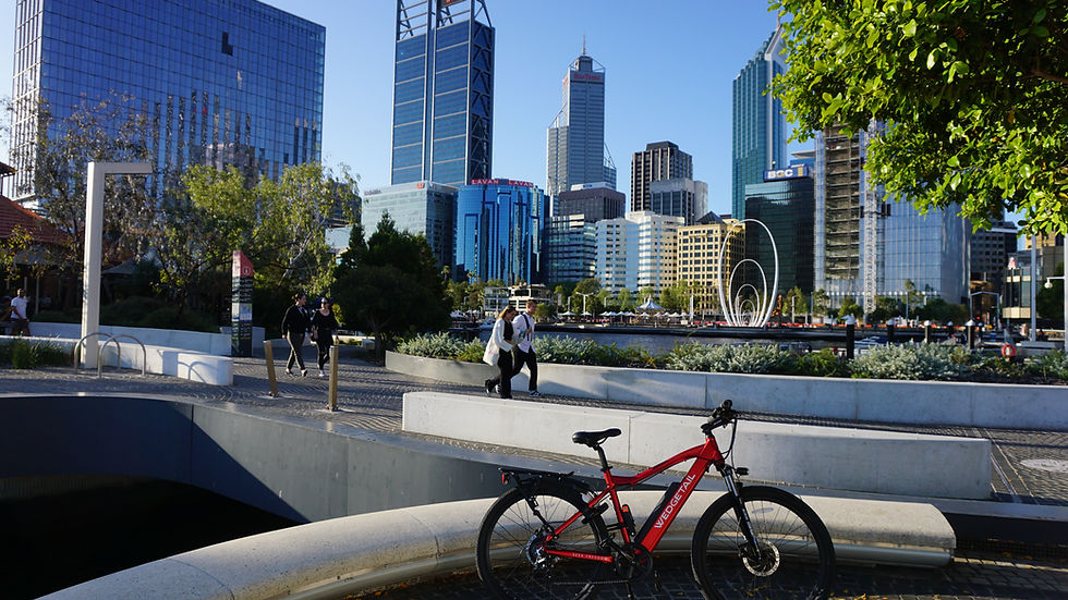 Matte Red WEDGETAIL ebike on a stone ledge in a sunny cityscape. Two people walk in the background. Skyscrapers and trees surround the area.
